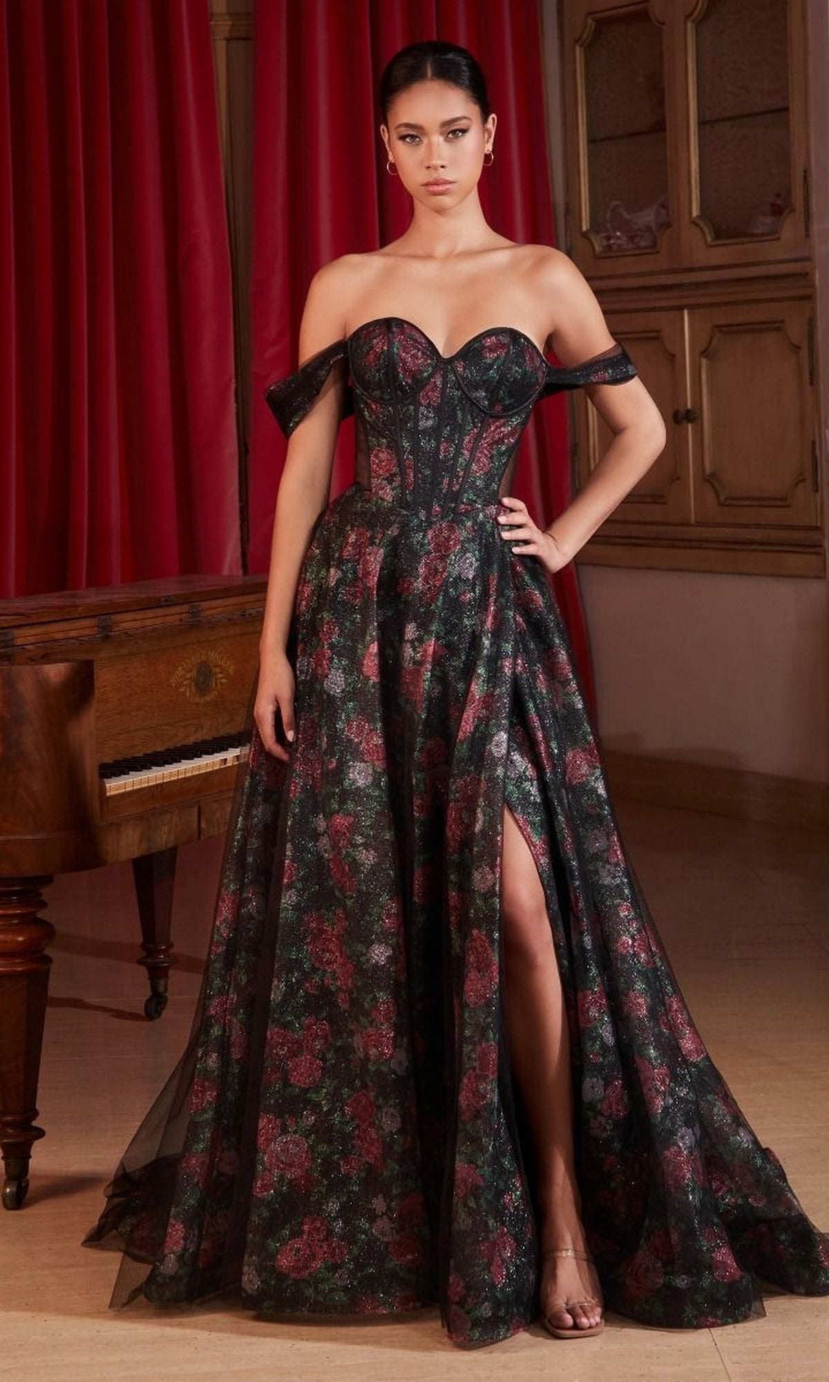 A woman poses indoors beside a wooden piano and red curtains, wearing the Off-Shoulder Long Floral-Print Prom Dress CD806, featuring black and red florals, a sheer overlay, and a high slit.